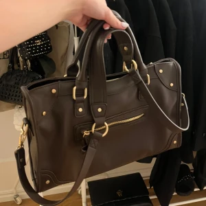 Väska med guld detaljer  - Snygg brun skinnväska med praktiska handtag och axelrem. Väskan har en dragkedja framtill och guldfärgade detaljer som ger en lyxig känsla. Perfekt för att bära med sig det nödvändigaste med stil. (Aldrig använd, nypris 499kr)