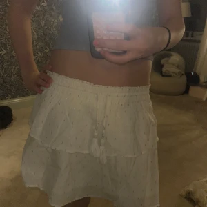 Vit volangkjol med knytband - Supersöt vit kortkjol med volanger och små glittriga detaljer. Kjolen har resår i midjan och dekorativa knytband med tofsar. Perfekt för sommarens alla tillfällen!