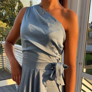 Ljusblå one shoulder långklänning från TFNC London - Säljer en elegant ljusblå långklänning från TFNC London. Klänningen har one shoulder-skärning, knytband i midjan och en slits framtill. Oanvänd med etikett kvar, storlek 8. 