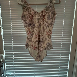Blommig baddräkt med volang - Säljer en söt baddräkt med blommigt mönster i vitt, lila och rött. Baddräkten har volangdetaljer vid axlarna och en feminin passform. Perfekt för sommarens bad och strandhäng! Hade den under graviditet till månad 7