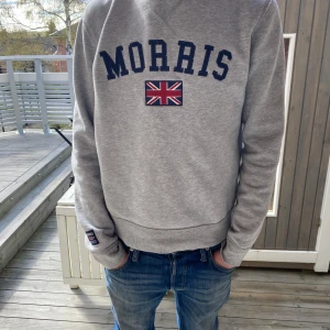 Grå sweatshirt från Morris - Säljer en grå sweatshirt från Morris med mörkblå text och brittisk flagga på bröstet. Tröjan har rund halsringning och långa ärmar. Perfekt för en avslappnad stil.