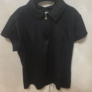 Svart kortärmad polotopp från Daily Women - Säljer en svart polotopp från Daily Women i storlek S. Toppen har korta ärmar, klassisk krage och dragkedja framtill med en liten dekorativ detalj. Perfekt för en stilren och enkel look.