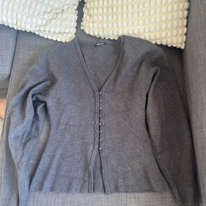 Mörkgrå kofta från Gina Tricot - Mörkgrå kofta från Gina Tricot med v-ringning och knappar framtill. Långärmad och i mjukt material, perfekt att slänga på sig över en topp. Enkel och stilren design som passar till det mesta. Storlek s