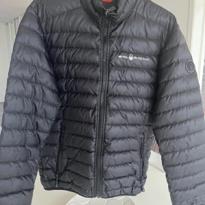 Svart pufferjacka från Sail Racing - Säljer en svart pufferjacka från Sail Racing med hög krage och dragkedja framtill. Jackan har quiltad design och två fickor med dragkedja. Perfekt för kyliga dagar och enkel att matcha med olika outfits.