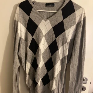 Grå argyle-mönstrad tröja från Massimo Dutti - Snygg grå tröja från Massimo Dutti med klassiskt argyle-mönster i svart, vitt och grått. V-ringad modell i 100% extrafint bomullsmaterial. Perfekt för en stilren och tidlös look.