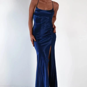 Marinblå långklänning i satin med slits - Säljer en marinblå långklänning i glansig satin med smala axelband och öppen rygg med snörning. Klänningen har en hög slits framtill och figurnära passform. Perfekt för bal eller festliga tillfällen.