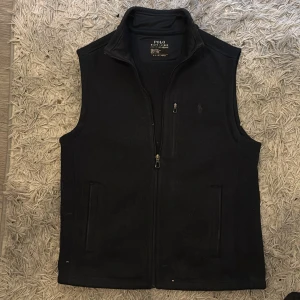  fleeceväst från Polo Ralph Lauren - Säljer en mötkblåf leeceväst från Polo Ralph Lauren med dragkedja framtill och tre fickor, varav en bröstficka med dragkedja. Perfekt att slänga över en hoodie eller tröja för extra värme.