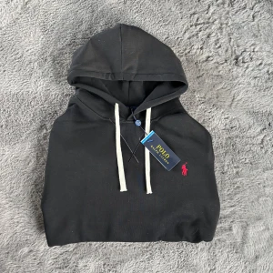 Svart hoodie från Polo Ralph Lauren - Säljer en svart hoodie från Polo Ralph Lauren med klassisk röd broderad logga på bröstet. Tröjan har huva med vita snören och en stor magficka framtill. Perfekt för en avslappnad och stilren look. Helt ny! Pris kan diskuteras vid snabb affär. Storlek M men sitter som S