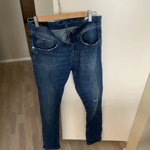 Mörkblå Dondup George - Säljer ett par mörkblå jeansbyxor med klassisk femficksdesign och diskreta slitningar på ben och bakficka (ej olycka). Byxorna har normal passform och raka ben. Perfekta till vardags för en avslappnad stil.