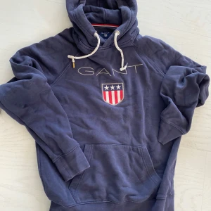 Mörkblå hoodie från GANT med broderad logga - Mörkblå hoodie från GANT med broderad logga på bröstet. Tröjan har huva med stor magficka. Perfekt för en avslappnad stil. Köparen står för frakt 🚚 