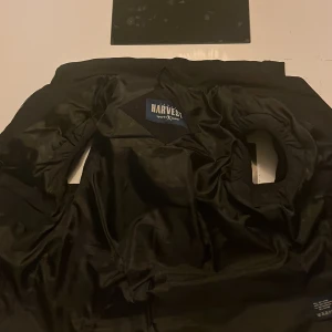 Svart dunväst från Harvex - Säljer en svart dunväst från Harvex med hög krage och dragkedja framtill. Västen har två sidofickor med dragkedja och är vadderad för extra värme. Perfekt för lager-på-lager under kyliga dagar.