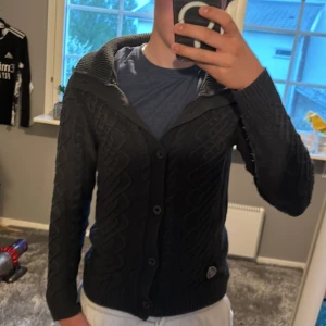 Mörkblå cardigan - Säljer en svart stickad kofta/cardiag med snyggt mönster och bred ribbad krage. Koftan har knappar framtill och en liten detalj på ena sidan nedtill. Perfekt för snygga dagar och passar till det mesta.