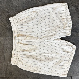 Vita linne shorts  - Snygga vita shorts med tunna svarta ränder och elastisk midja med snörning. Shortsen har sidofickor och är tillverkade i ett lätt och luftigt tyg, perfekt för varma dagar.