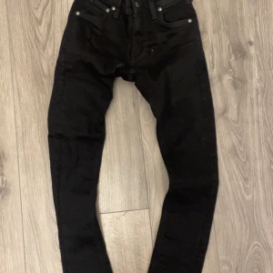 Svarta jeans från Nudie Jeans - Säljer ett par svarta jeans från Nudie Jeans med klassisk femficksdesign och smal passform. Jeansen har svarta sömmar och subtila detaljer på bakfickorna. Perfekta för dig som gillar en stilren look.