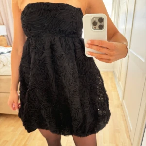 Svart axelbandslös kortklänning med rosmönster - Säljer en svart axelbandslös kortklänning med struktur i form av rosor över hela tyget. Klänningen har en voluminös kjol och sitter åt upptill. Perfekt för dig som vill sticka ut med en unik och elegant look❣️❣️prislapp kvar! Nypris 600kr