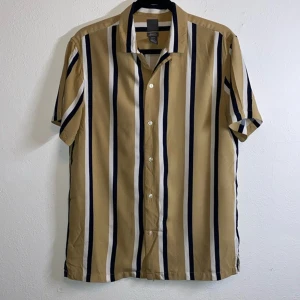 Beige och vit randig kortärmad skjorta - Snygg kortärmad skjorta med vertikala ränder i beige, vitt och mörkblått. Skjortan har klassisk krage och knäppning framtill. Perfekt för en avslappnad och stilren look.