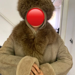 Lång beige pälsjacka från AmoreSS - Säljer en lång beige kappa från AmoreSS med bred pälskrage och pälskantade ärmslut. Jackan har en klassisk rak passform och stängs med knappar framtill. Perfekt för kalla dagar när du vill vara både varm och snygg.
