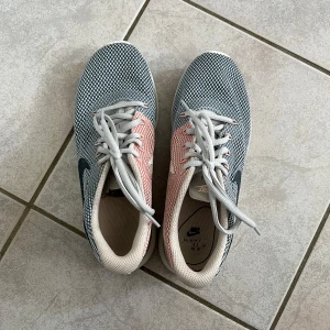 Nike sneakers i grått och rosa - Säljer ett par Nike sneakers i grått och rosa med vit sula och vit Nike-logga på sidan. Skorna har snörning och är tillverkade i syntetmaterial med mesh för bra andningsförmåga. Perfekta för vardagsbruk eller träning. Storlek 38, varsamt använda 💗