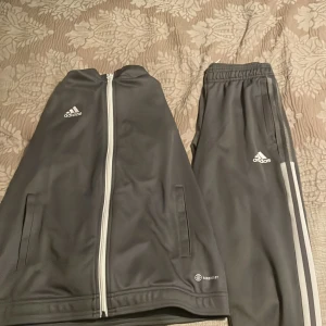 Grå Adidas träningsbyxor med vita ränder - Säljer ett par grå Adidas träningsbyxor med klassiska vita ränder längs sidorna och Adidas-logga på låret. Byxorna har resår i midjan och är perfekta för sport eller chill. Matchande jacka finns också.