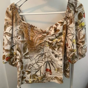 Blommig blus från H&M - Säljer en blommig blus från H&M i storlek 36. Toppen har puffiga trekvartsärmar, v-ringning och ett tropiskt mönster i beige, brunt, grönt och inslag av rött och gult. Tillverkad i linneblandning och perfekt för sommaren. Aldrig använd 