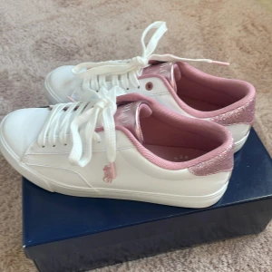 Vita sneakers från Polo Ralph Lauren med rosa detaljer - Säljer ett par fräscha vita sneakers från Polo Ralph Lauren med rosa insida och glittriga rosa detaljer vid hälen. Klassisk design med snörning och Polo-logga på sidan. Är i stl 39 men passar till 38. Säljer då de är försmå.
