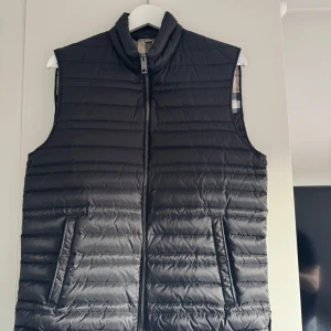 Svart dunväst Burberry gåsdun  - Snygg svart dunväst från Burberry med klassiskt rutigt mönster på insidan. Västen har två fickor med knappstängning på utsidan och en ficka på insidan. Kvitto finns! Perfekt för en stilren look. 48 är medium men skulle säga att den passar någon som har S eller M. Mvh Oliver
