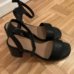 ASOS skor med klack och remmar - Säljer ett par stilrena svarta skor med bred klack och justerbara remmar runt vristen. Skorna har öppen tå och bred rem över foten, vilket ger en modern och enkel look. Fint skick, bara testade inomhus. 