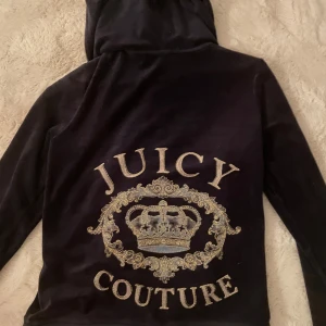 Mörkblå velour hoodie från Juicy Couture - Mjuk och mörkblå hoodie i velour från Juicy Couture med dragkedja framtill och stor broderad krona samt logga i guld på ryggen. Huva och fickor framtill.
