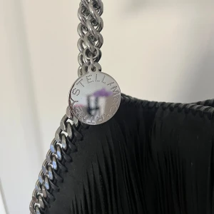 Svart axelväska med fransar från Stella McCartney - Säljer en svart axelväska från Stella McCartney med långa fransar och kedjedetaljer. Väskan har en rund metallbricka med logga och dragkedja upptill. Insidan är fodrad i ljusrosa tyg med logotypmönster. Äktahetskontrollerad och har alla koder! Nypris runt 13 000kr