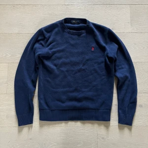 Mörkblå stickad tröja från Polo Ralph Lauren - Säljer en klassisk mörkblå stickad tröja från Polo Ralph Lauren med rund halsringning och röd broderad logga på bröstet. Tröjan har ribbade muddar vid ärmslut och nederkant. Perfekt för en stilren look! Säljer pga att den är förliten