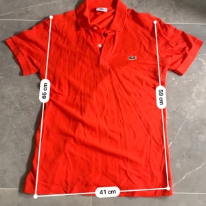 Orange piké t-shirt från Lacoste - Säljer en orange piké t-shirt från Lacoste i slim fit-modell. Klassisk design med krage, två knappar och den ikoniska Lacosteloggan broderad på bröstet. Perfekt för dig som gillar stilrena och sportiga plagg.passar perfekt nu till sommaren!