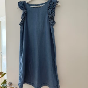 Blå ärmlös klänning med volang från Mango - Ärmlös klänning i denimlook med volangdetaljer vid axlarna och rund halsringning. Perfekt för sommardagar och enkel att styla med sneakers eller sandaler. Storlek S. 78 cm lång. Sparsamt använd(2-3 gånger), mycket fint skick. Tvättad. Djurfritt och rökfritt hem.