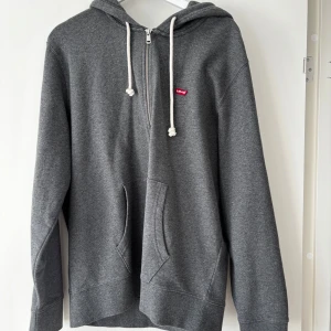 Grå hoodie med dragkedja från Levi's - Säljer en grå hoodie från Levi's i storlek L. Tröjan har dragkedja framtill, huva med vita snören och två fickor. Perfekt för en avslappnad stil och enkel att matcha med det mesta.