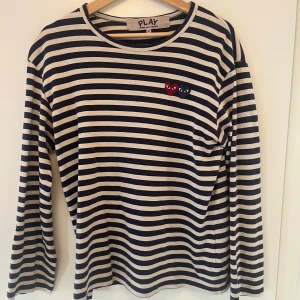 Randig långärmad tröja från Comme des Garçons Play - Säljer en klassisk långärmad tröja från Comme des Garçons Play med marinblå och vita ränder. Tröjan har ett broderat hjärta på bröstet och rund halsringning. Perfekt för en stilren och avslappnad look.