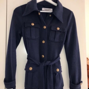 Mörkblå tröja med bälte och guldknappar - Snygg mörkblå tröja från Cristina Gavioli med markerad krage, långa ärmar och bälte i midjan. Tröjan har dekorativa guldknappar framtill och på fickorna, samt fyra fickor för en cool utility-look.