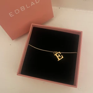 Guldigt halsband med bokstaven E - Säljer ett stilrent halsband i guld med bokstaven E Perfekt som personlig accessoar eller present. Kedjan är tunn och elegant, och halsbandet levereras i en edblad-ask (halsbandet är inte från edblad)