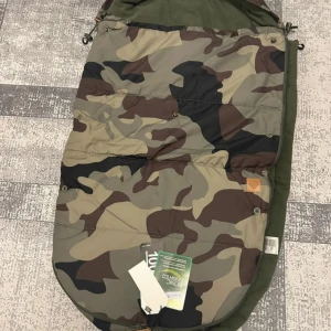 Kameramönstrad åkpåse - Säljer en åkpåse med kameramönster i gröna och bruna nyanser. Utsidan är mönstrad medan insidan är enfärgat grön och mjuk. Passar perfekt för kalla dagar och har flera öppningar för sele.
