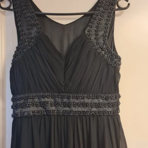 Svart långklänning med pärldetaljer - Elegant svart långklänning med pärldetaljer längs axelbanden och midjan. Klänningen har v-ringning med transparent mesh upptill och ett mjukt, fallande tyg. Perfekt för festliga tillfällen..Om det behövs kan jag skicka bättre bilder på klänningen vid intresse 