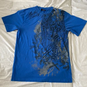 Blå t-shirt med tryck från Southpole - Säljer en blå t-shirt från Southpole med stort svart grafiskt tryck och textdetaljer på framsidan. T-shirten har rund hals och är kortärmad. Tillverkad i 100% bomull, perfekt för en cool och avslappnad stil.