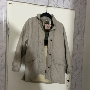 Beige quiltad jacka från KappAhl - Snygg beige quiltad jacka från KappAhl med rutigt innerfoder. Jackan har stora fickor framtill, knappar vid ärmslut och hög krage. Perfekt för kyligare dagar och enkel att matcha med olika outfits.