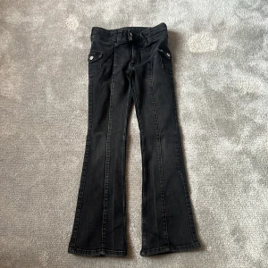 Svarta bootcut jeans från H&M - Svarta jeans från H&M med bootcut-ben och låg midja. Byxorna har justerbar midja, två framfickor med lock och knapp samt klassisk femficksdesign. Snygga kontrastsömmar och silverfärgade knappar framtill.
