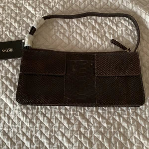 Brun clutch/axelväska från BOSS med ormmönster och ormskinn  - Säljer en brun clutch/axelväska från BOSS med snyggt ormmönstrat mönster och dragkedja upptill. Väskan har en smal axelrem och stilren design, perfekt för att bära det viktigaste. Märkets tagg sitter kvar. Väskan är helt ny som du ser på tagen och plasten. 