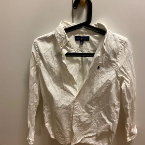 Vit skjorta från Ralph Lauren - Klassisk vit skjorta från Ralph Lauren med broderad logga på bröstet. Skjortan har lång ärm, knappar framtill och krage. Perfekt för en clean och stilren look.