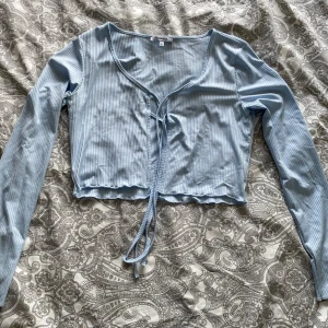 Ljusblå ribbad croppad topp från Chiquelle - Säljer en ljusblå croppad topp från Chiquelle med långa ärmar och knyt framtill. Toppen är ribbad och har en v-ringad halsringning. Perfekt för att skapa en trendig och avslappnad look.