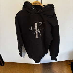 Svart hoodie från Calvin Klein - Svart hoodie från Calvin Klein med stor glittrig CK-logga framtill. Klassisk känguruficka och huva med dragsko. Perfekt för en avslappnad och trendig stil. 