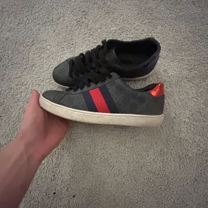 Gucci sneakers med röd och blå rand - Priset är inte fast, Säljer ett par Gucci sneakers i svart med monogrammönster, röda och blå ränder på sidan samt metallic-detaljer i rött och lila på hälen. Skorna har snörning och vit platt sula. Skorna är helt äkta men har dock inget äktehetsbevis, finns tyvärr heller inget till skorna 