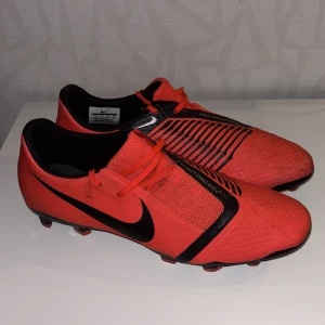 Nike Phantom Vision fotbollsskor i rött och svart - Säljer ett par Nike Phantom Vision fotbollsskor i rött med svarta detaljer och snörning. Skorna har en låg profil och mönstrad ovandel för bättre bollkontroll. Perfekta för spel på gräsplaner.
