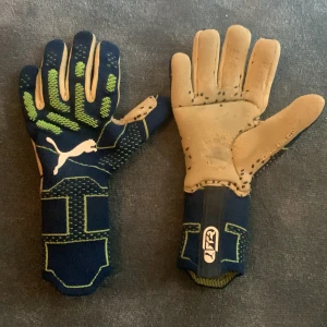 Blåa och beige fotbollshandskar från Puma - Säljer ett par blåa fotbollshandskar från Puma med beige insida och gröna detaljer på ovansidan. Handskarna har lång mudd och är designade för bra grepp och skydd vid spel. Målvaktshandskarna är inte i ett särskilt bra skick, därför säljer jag dem för ett mycket billigare pris än vad jag köpte dem för. Pris är diskuterbart. 