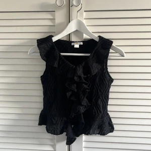 Svart volangblus med struktur - Säljer en svart ärmlös blus med volangdetaljer framtill och vågig struktur i tyget. Blusen har v-ringning och en lätt croppad passform som ger en trendig look.