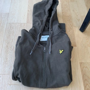 Olivgrön hoodie med dragkedja från Lyle & Scott - Säljer en olivgrön hoodie från Lyle & Scott med dragkedja framtill och klassisk gul logga på bröstet. Tröjan har huva med dragsko och två fickor framtill. Perfekt för en avslappnad stil.